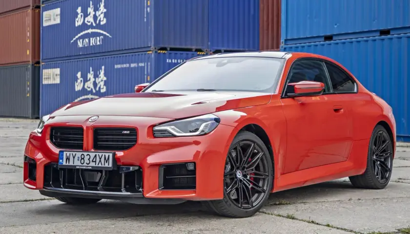 Pomarańczowe BMW M2 Competition z czarnymi felgami stoi na tle niebieskich kontenerów.