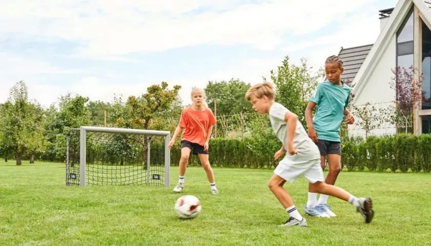 Drei Kinder üben im Garten Fußball. Ein Junge dribbelt, während ein Mädchen und ein weiterer Junge zuschauen.