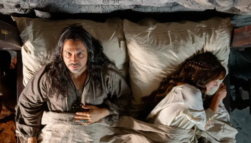 Un homme aux longs cheveux regarde la caméra, allongé dans un lit à côté d'une femme endormie. Un air de mystère plane sur ce moment, rappelant les intrigues de Mordred dans Kaamelott.