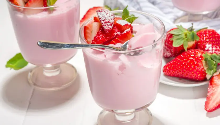 Różowa panna cotta z truskawkami, udekorowana listkami mięty i posypana cukrem pudrem. Obok świeże truskawki.