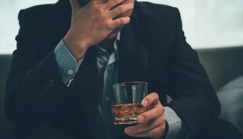 Mężczyzna w garniturze z ręką na czole, trzymający szklankę z bursztynowym płynem. Może to symbolizować problemy, stres lub nawet renta alkoholowa.