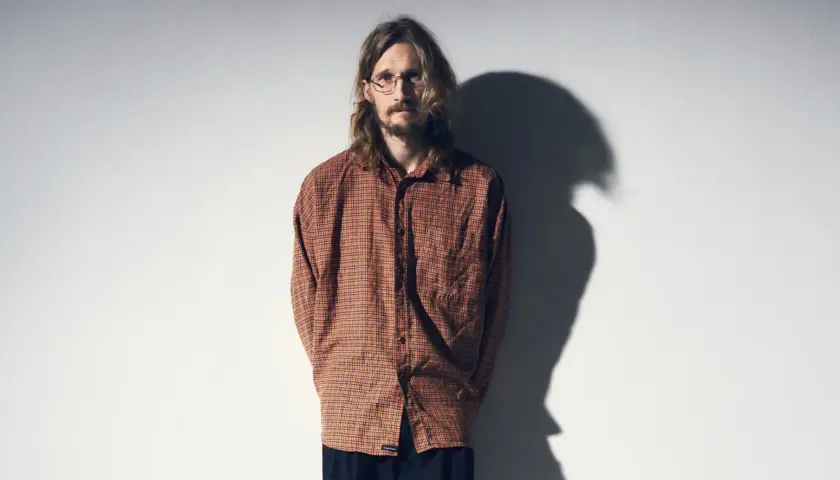Marcus Berggren komiker, med sitt karaktäristiska hår och glasögon, står framför en vit vägg med en skugga som dansar bakom honom.