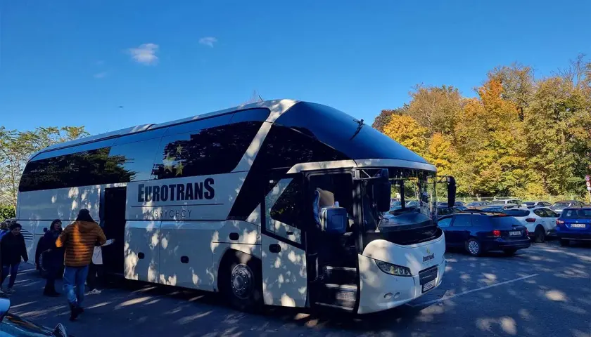 Biały autokar firmy Eurotrans z otwartymi drzwiami, zaparkowany na tle jesiennych drzew i innych samochodów.