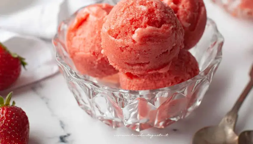 Coppa di sorbetto cremoso alle fragole, fresca e invitante, con fragole fresche e cucchiaini d'argento su sfondo marmorizzato.