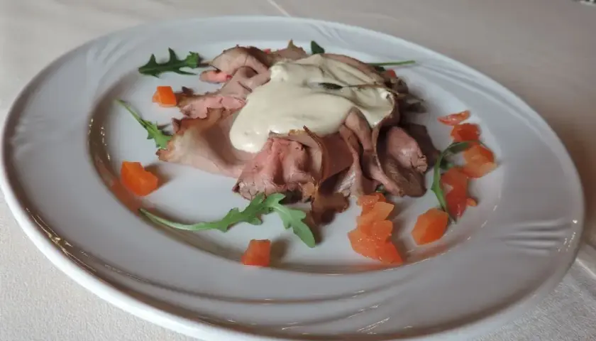 Vitello tonnato, un classico con origini piemontesi, servito su un piatto bianco con pomodorini e rucola.