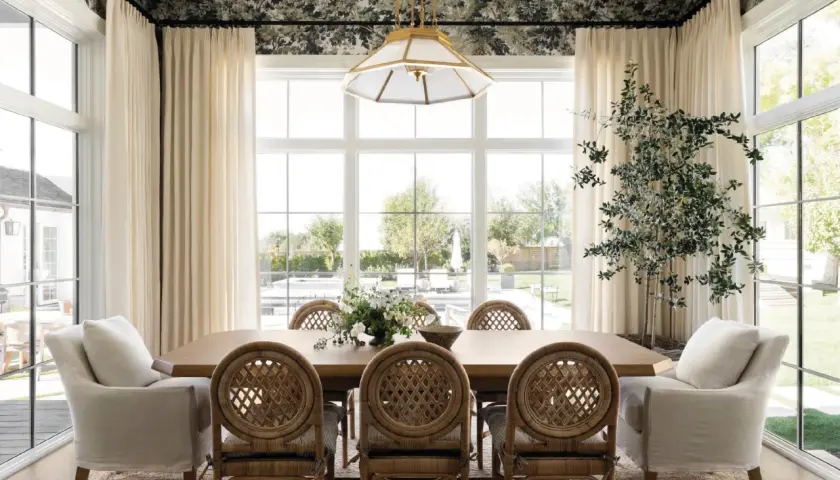 A bright dining room in a Studio McGee home features a large wooden table with rattan chairs, plush armchairs, and a floral wallpaper.