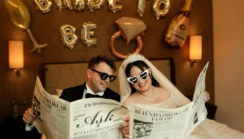 Para Młoda czyta gazetę "The Wedding Post" o miłości, przygotowując się do wesela krok po kroku. Balony "Bride to Be" i kieliszki tworzą atmosferę.