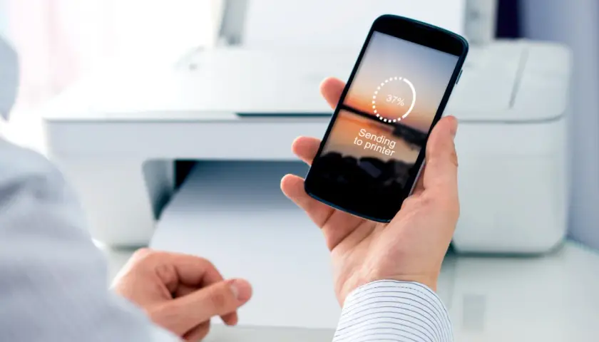 Dłoń trzyma smartfon z napisem "Sending to printer", pokazując jak drukować z telefonu. W tle biała drukarka.