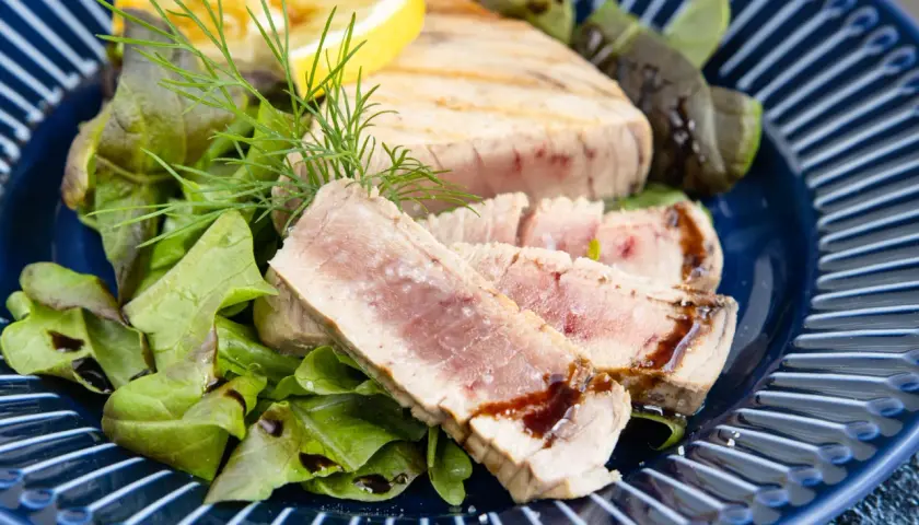 Saftiges Thunfisch Steak, medium gebraten, serviert mit frischem Salat, Dill und einer Zitronenspalte auf einem blauen Teller.