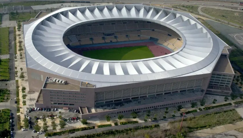 Imponujące, okrągłe **Estadio La Cartuja** z charakterystycznym, falującym dachem, otoczone zielenią i drogami.