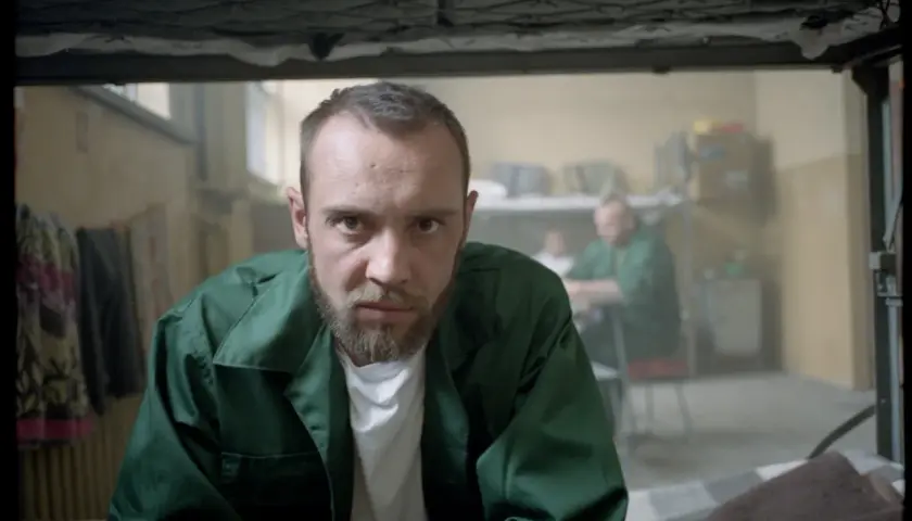 Mężczyzna w zielonej kurtce, jak z filmu "Jak zostałem gangsterem. Historia prawdziwa", patrzy wprost. W tle inni więźniowie.