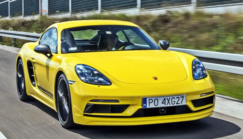 Żółty Porsche Cayman T pędzi po drodze, kierowca w czapce z daszkiem za kierownicą.