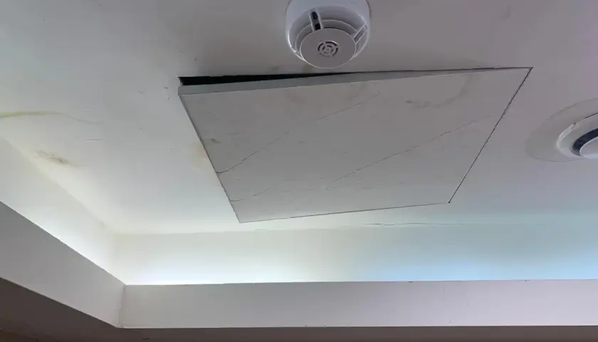 Un soffitto in cartongesso con una botola aperta, un rilevatore di fumo e macchie d'acqua. Il montaggio botola cartongesso è visibile.