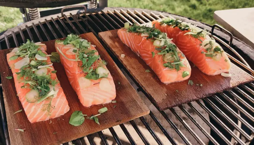 Quatre pavés de saumon sur planche de cèdre, garnis d'herbes fraîches et d'ail, prêts à être grillés.