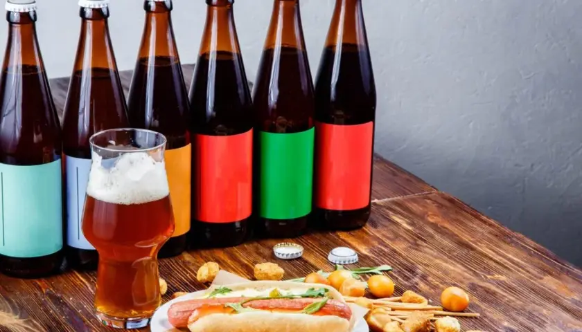 Bouteilles de bière espagnole alignées, un verre de bière ambrée et un hot-dog garni sur une table en bois.