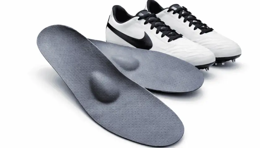 Zwei orthopädische fußball einlagen liegen vor einem Paar weißer Fußballschuhe mit schwarzen Schnürsenkeln und Swoosh-Logo.