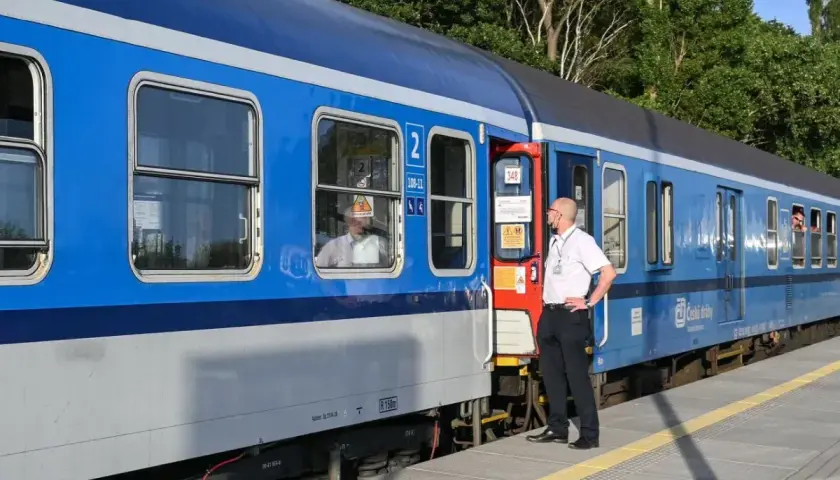 Pracownik kolei stoi przy drzwiach niebieskiego pociągu. Pasażerowie wyglądają przez okna. Czy można kupić bilet w pociągu?