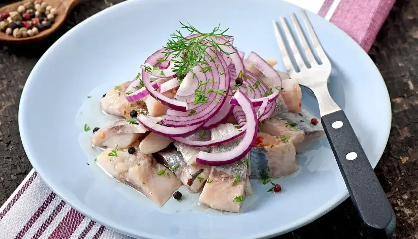 Kawałki śledzia z cebulą i koperkiem na jasnoniebieskim talerzu, obok widelec.