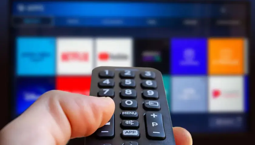 Hand håller fjärrkontroll framför en TV-skärm med app-ikoner. Kanske dags att välja en film eller en dokumentär om ze rappare.