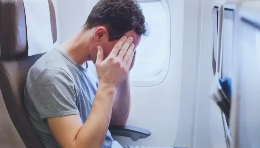 Mężczyzna w samolocie z rękami na twarzy, wyraźnie cierpiący na jet lag po długiej podróży.