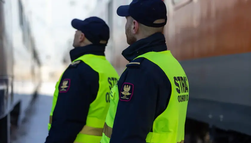 Dwóch funkcjonariuszy straży ochrony kolei w kamizelkach odblaskowych stoi przy pociągu, czuwając nad bezpieczeństwem.