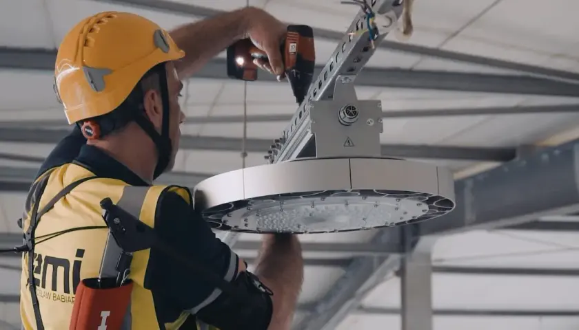 Pracownik w żółtym kasku i kamizelce montuje nowoczesne oświetlenie LED. To wymiana oświetlenia na LED – remont czy modernizacja hali.