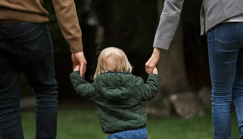 Dziecko z blond włosami, w zielonej kurtce, trzyma za ręce rodziców. To obraz przysposobienia niepełne, pełnego miłości i nadziei.
