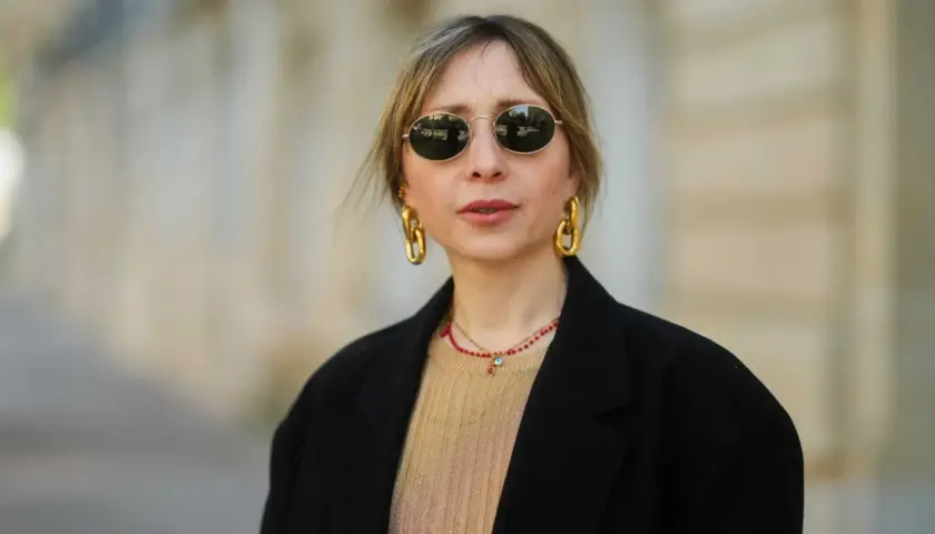 Frau mit stylischer curtain bangs frisur, runden Sonnenbrillen und auffälligem Goldschmuck.