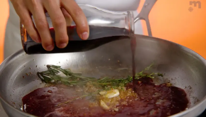 Une main verse du jus de viande dans une poêle avec de l'ail et des herbes.