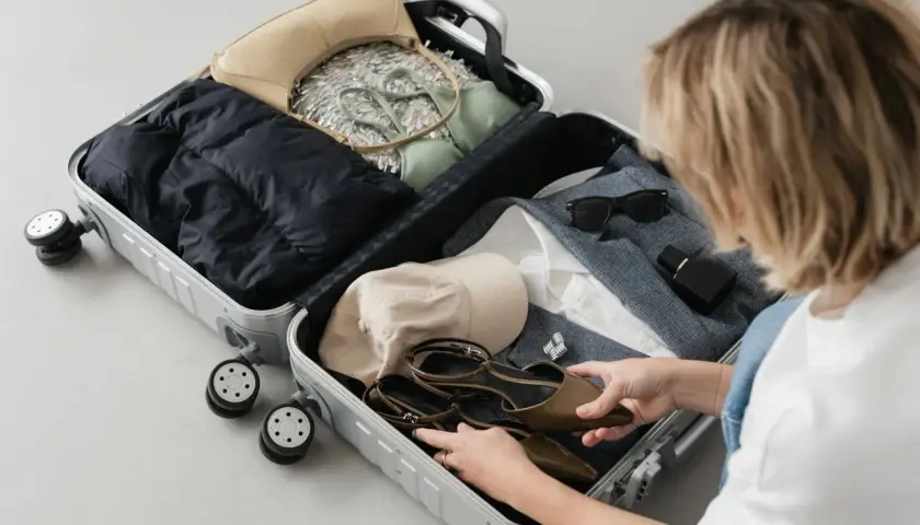 Kobieta pakuje do walizki: czarną kurtkę, zielony komplet, beżową torebkę, czapkę, buty, okulary i perfumy. Gotowa na podróż z enter air bagaż.