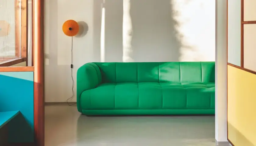 Ein leuchtend grünes Sofa steht vor einer weißen Wand, an der eine orangefarbene Lampe hängt. Sonnenlicht wirft Schatten. Mette hay.