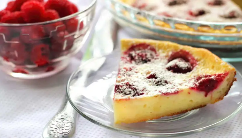 Une part de clafoutis framboise, saupoudrée de sucre glace, à côté d'un bol de framboises fraîches.