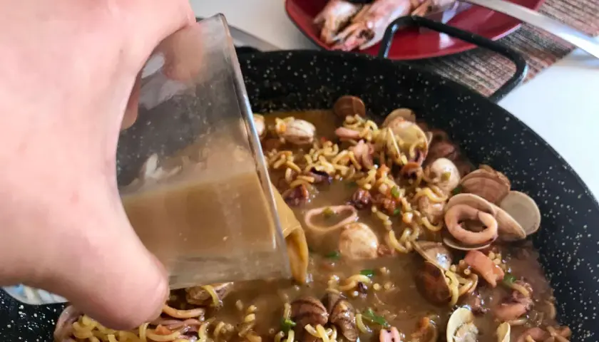 Vertiendo caldo en una paellera con fideos, calamares y almejas. Así se hace fideuá.