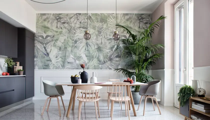 Un soggiorno moderno con carta da parati a tema tropicale, un tavolo da pranzo con sedie in legno e un'elegante cucina grigia. Chi mette la carta da parati sceglie un tocco di natura.