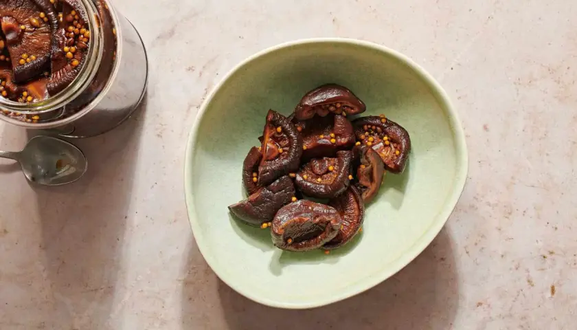 A jar and bowl of glistening pickled shiitake mushrooms, studded with mustard seeds.