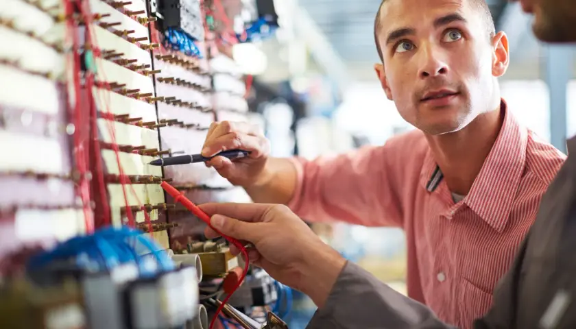 Dwóch mężczyzn pracuje przy panelu elektrycznym, jeden trzyma miernik.