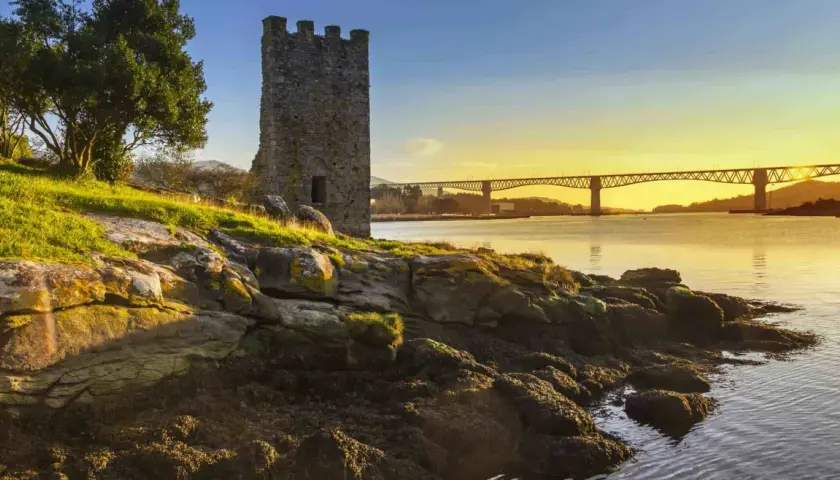 Torre medieval junto a un río al atardecer, con un puente al fondo. Imagina el tren Madrid Ferrol pasando por allí.