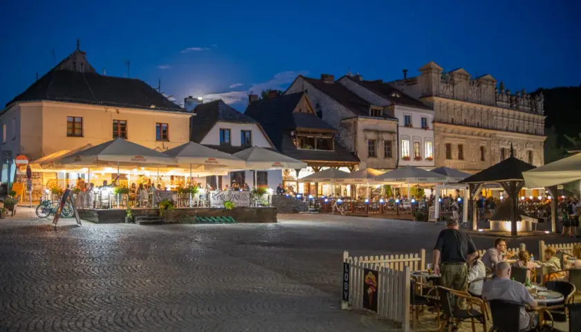 Wieczorny rynek w Kazimierzu Dolnym tętni życiem. Ludzie siedzą przy stolikach pod parasolami, delektując się posiłkami i atmosferą.