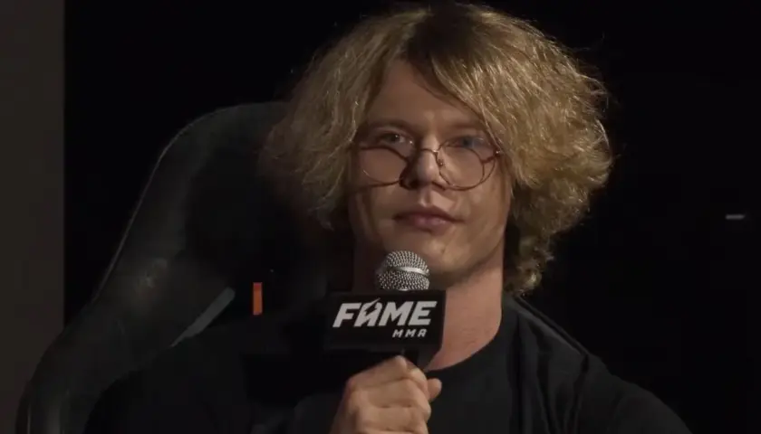 Natan Marcon z burzą blond włosów i okularami, trzymający mikrofon FAME MMA. Jego wiek jest trudny do określenia, ale wygląda na młodego mężczyznę.