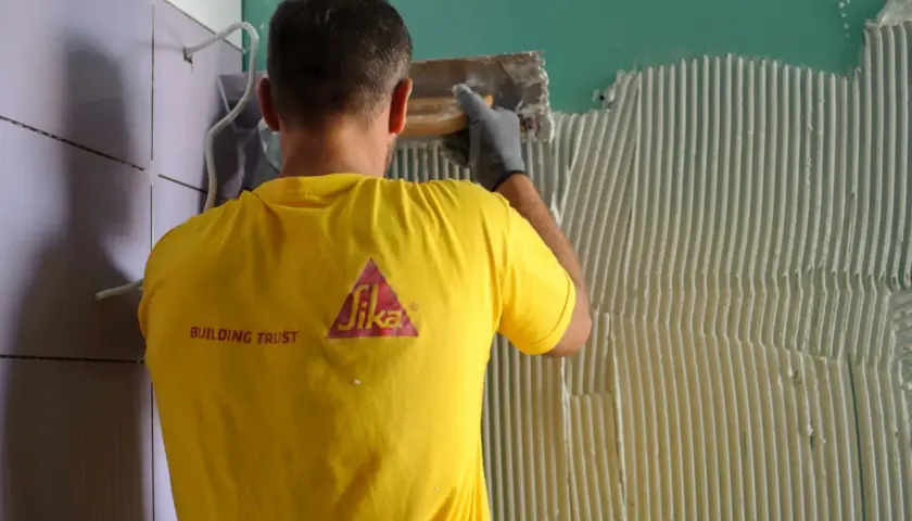 Operaio con maglia gialla Sika applica colla per la partenza posa piastrelle a muro.