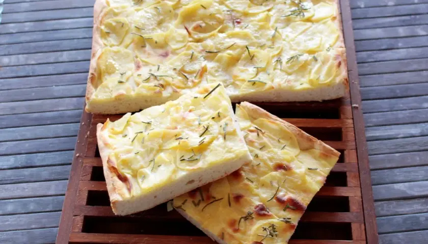 Focaccia con lievito istantaneo, ricoperta di patate sottili e rosmarino, tagliata a fette su un tagliere di legno.