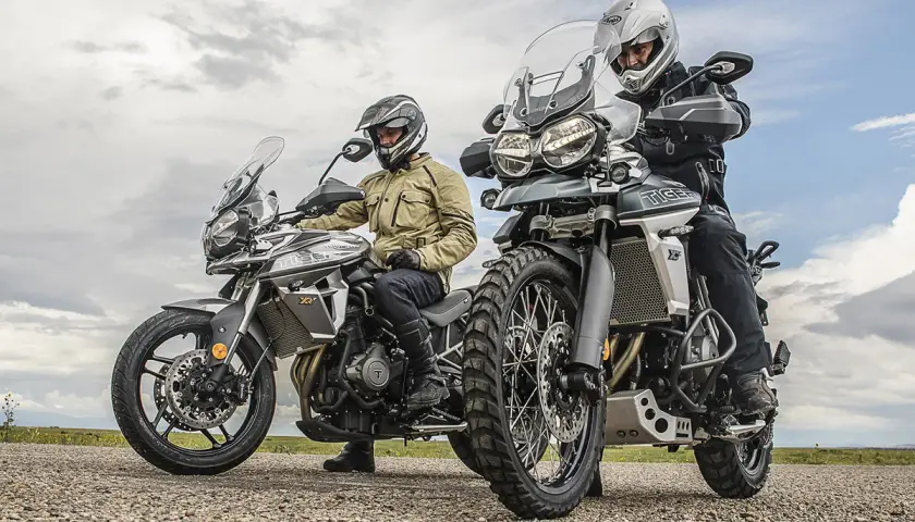 Dos motociclistas con cascos y equipo de aventura listos para explorar caminos y **trail asfalticas** bajo un cielo nublado.
