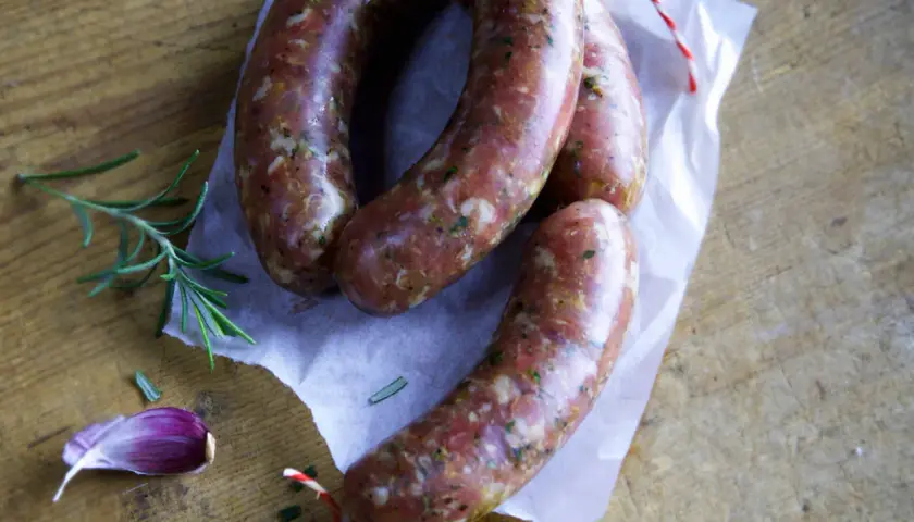 Frische italienische Bratwurst Salsiccia, bereit für ein köstliches Rezept. Rosmarin und Knoblauch liegen daneben.