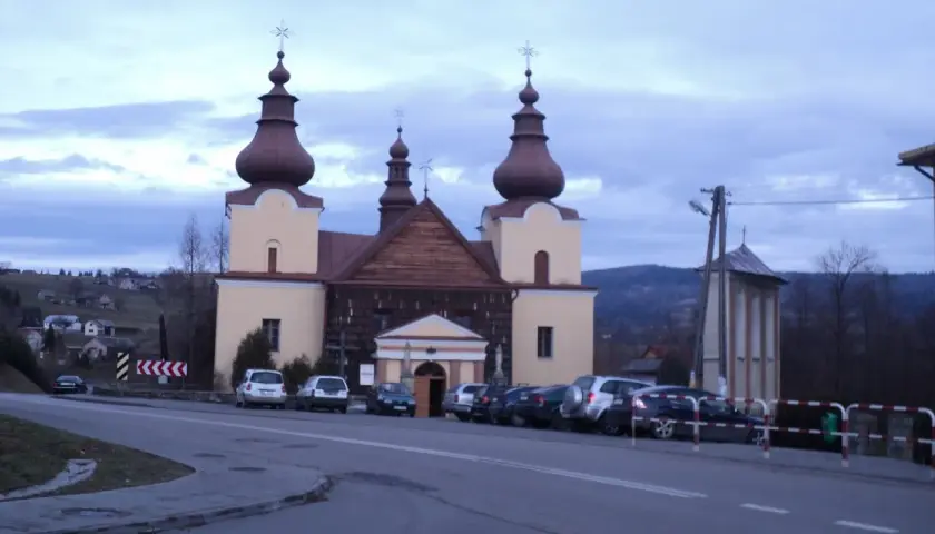 Cerkiew łemkowska z charakterystycznymi kopułami, otoczona samochodami, na tle pagórkowatego krajobrazu.