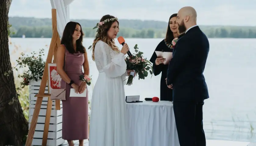 Panna młoda z mikrofonem i bukietem mówi podczas ceremonii. Obok stoi pan młody, urzędniczka i świadkowa. To ślub jednostronny nad jeziorem.