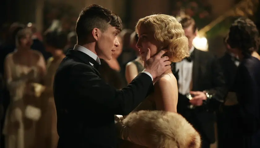 Tommy Shelby czule dotyka policzka Grace, jednej z kluczowych kobiet w świecie Peaky Blinders. W tle widać innych gości balu.