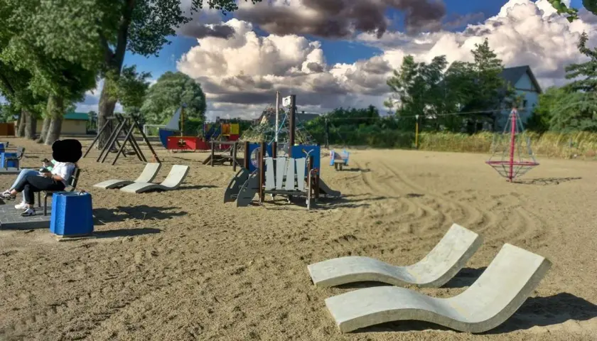 Pusta lubczyna plaża z placem zabaw i leżakami. Kobieta siedzi na ławce, patrząc na telefon.