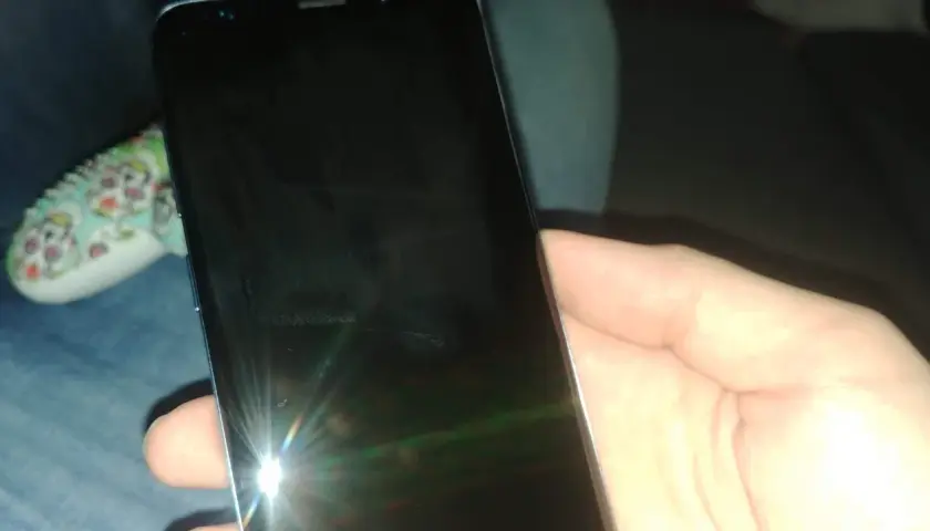 Dłoń trzyma czarny telefon, na którego ekranie widać odbicie światła i ledwo widoczną kreskę na telefonie.