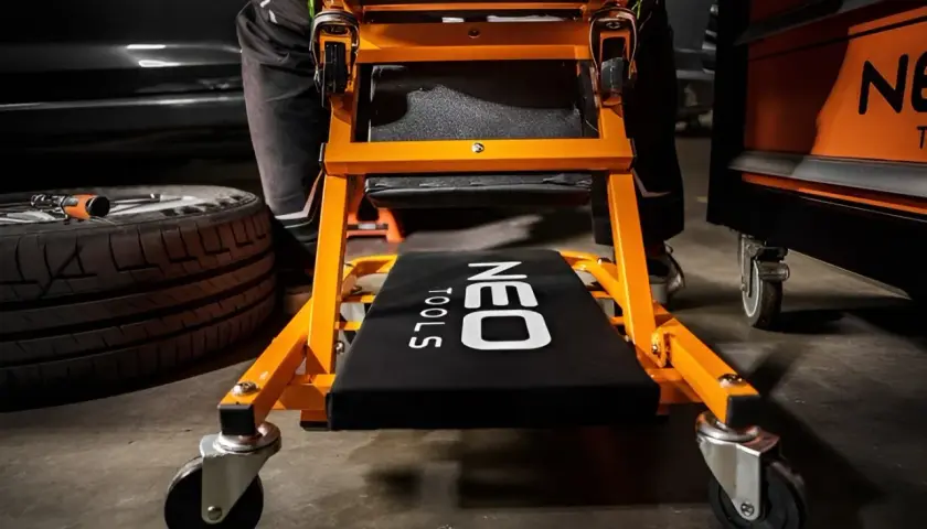 Pomarańczowa leżanka pod auto z kółkami, gotowa do pracy. Obok leży opona.