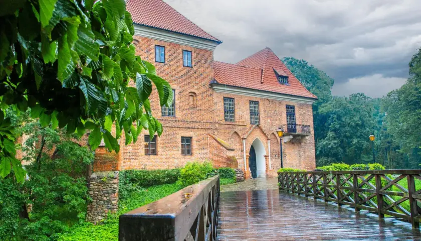 Deszczowy dzień nad zamkiem w Oporowie. Drewniany mostek prowadzi do ceglanego budynku, otoczonego zielenią.